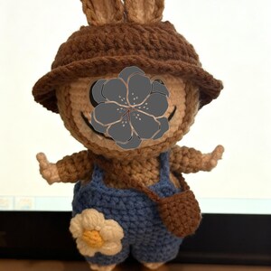 Peut inclure: Poup&eacute;e lapin au crochet faite &agrave; la main. Le lapin porte un chapeau marron, une salopette bleue et un petit sac marron. La salopette a une fleur blanche et jaune. Le lapin est fait de fil marron et beige.