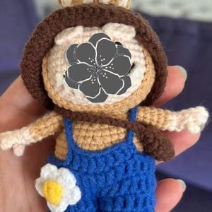 Peut inclure: Poup&eacute;e au crochet faite &agrave; la main repr&eacute;sentant un personnage de lapin. La poup&eacute;e porte une salopette bleue avec une fleur blanche et jaune. Elle a un chapeau marron et un petit sac marron. La poup&eacute;e est faite de fil beige, marron et blanc.