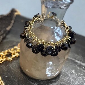 May include: A gold-toned chain bracelet adorned with black buttons and faceted beads. The bracelet is draped over a clear glass vase, with a gold chain visible in the background. The bracelet's design is unique and eye-catching.