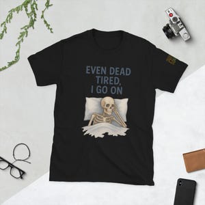 Puede incluir: Camiseta negra con el texto "EVEN DEAD TIRED, I GO ON" y un dibujo de un esqueleto en la cama. El esqueleto es pálido, con una mano en la cabeza. La camiseta tiene un diseño de manga corta.