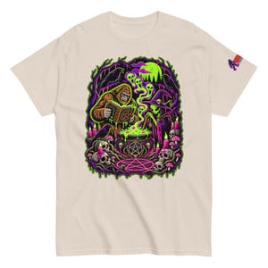 May include: A beige t-shirt featuring a graphic print of a brown sasquatch and a green alien standing over a bubbling cauldron. The scene includes skulls, candles, a pentagram, and ravens in a dark forest setting. A small logo is on the sleeve.
