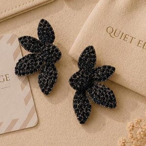 May include: A pair of black flower-shaped earrings with small, dark gemstones. The earrings are displayed on a beige surface, next to a small beige pouch and a card with the text "QUIET EDGE". The earrings are a jewelry item.