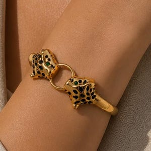 May include: A gold-toned metal cuff bracelet featuring two leopard head motifs facing each other. The leopard heads are detailed with black enamel spots and small green gemstone eyes, connected by a central gold ring on the open-ended band.