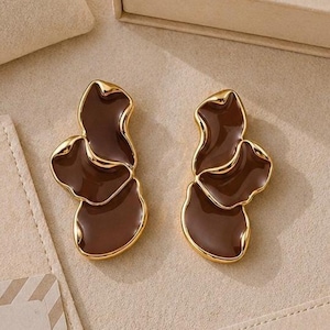 May include: A pair of brown and gold earrings with a floral design. The earrings are displayed with a beige jewelry bag, box, and cards. The text "QUIET EDGE" and "GIFT READY" are visible.