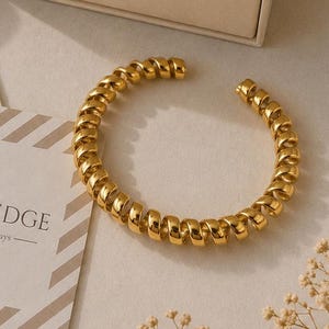 Puede incluir: Una pulsera de brazalete en espiral de tono dorado junto a una bolsa de tela beige con cordón, un joyero beige a juego y una tarjeta cuadrada con rayas diagonales. La bolsa, la caja y la tarjeta tienen impreso el texto Quiet Edge.