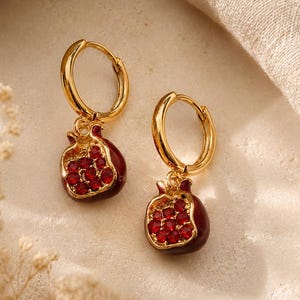 May include: A pair of gold-coloured hoop earrings. Each earring has a pendant shaped like a pomegranate, with a dark red enamel exterior and a cluster of red gemstones representing the seeds. The earrings are displayed on a neutral-toned surface.