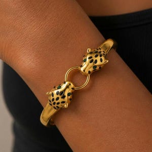 May include: A gold-toned bangle bracelet featuring two stylised leopard heads facing each other, connected by a central gold ring. The leopard heads are decorated with black enamel spots and small green accents for the eyes, creating a bold design.