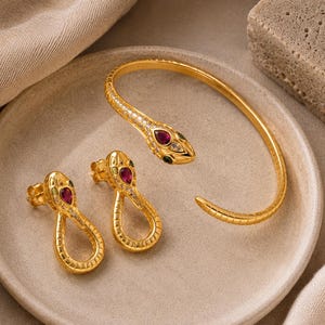 May include: A gold-toned jewelry set consisting of a snake-shaped cuff bracelet and two matching snake-shaped stud earrings. Each piece features a textured scale pattern, a teardrop-shaped red stone on the head, and small clear accent stones.
