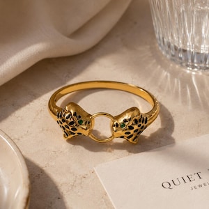 Puede incluir: Un brazalete de metal dorado con dos motivos de cabeza de leopardo enfrentados, unidos por un pequeño anillo circular. Las cabezas de leopardo tienen manchas de esmalte negro y pequeños ojos de piedra verde. Una tarjeta cercana dice QUIET EDGE JEWELLERY.