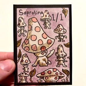 Puede incluir: Una tarjeta dibujada a mano con un borde negro que presenta criaturas con forma de hongo. La tarjeta tiene un fondo rosa con la palabra "Saproling" y el número "1/1" en la parte superior. Los hongos son beige con lunares rosas.