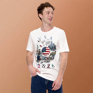 Pu&ograve; includere: T-shirt bianca con grafica colorata. Il design include la scritta "USA", la bandiera americana, una mappa del mondo e monumenti. Il numero "2024" e un pallone da calcio sono in basso. La t-shirt &egrave; indossata da una persona.