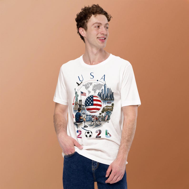 Pu&ograve; includere: T-shirt bianca con grafica colorata. Il design include la scritta "USA", la bandiera americana, una mappa del mondo e monumenti. Il numero "2024" e un pallone da calcio sono in basso. La t-shirt &egrave; indossata da una persona.