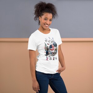 Pu&ograve; includere: T-shirt bianca con un design grafico colorato con la scritta "USA" e "2026". Il design include la bandiera americana, un pallone da calcio e illustrazioni di monumenti. La maglietta &egrave; realizzata in un materiale morbido.