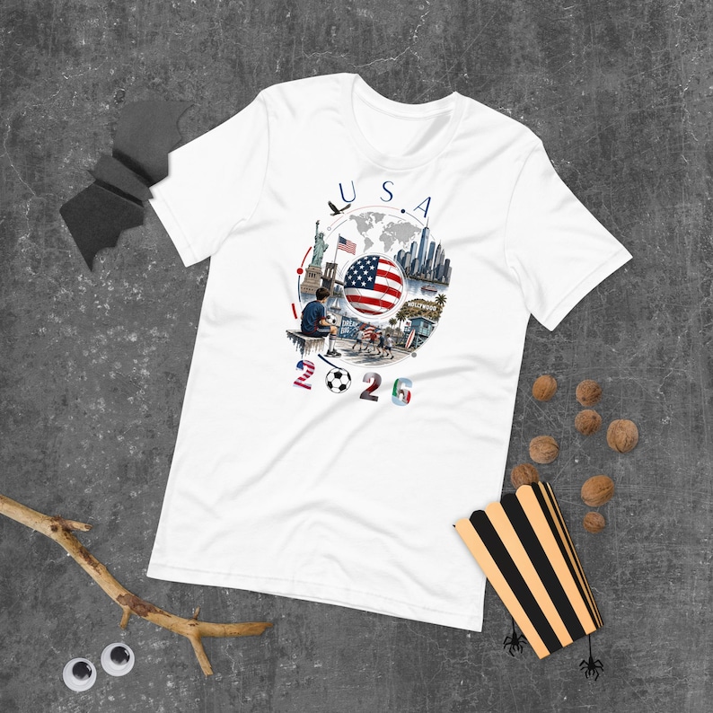 Pu&ograve; includere: T-shirt bianca con un design grafico con la scritta "USA 2026" e un pallone da calcio. Il design include la bandiera americana, una mappa del mondo e illustrazioni di monumenti.