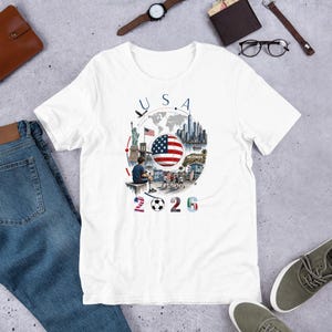 Pu&ograve; includere: T-shirt bianca con un design grafico colorato con la scritta "USA 2026". Il design include la bandiera americana, un pallone da calcio e illustrazioni di monumenti come la Statua della Libert&agrave; e l'insegna di Hollywood.