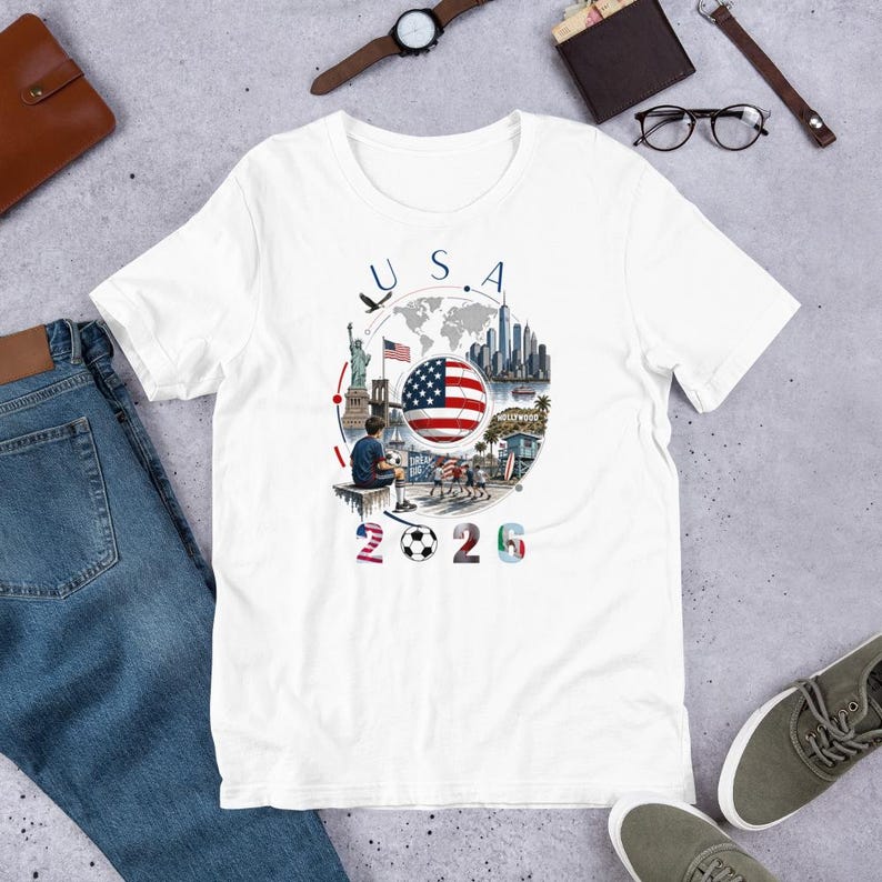 Pu&ograve; includere: T-shirt bianca con un design grafico colorato con la scritta "USA 2026". Il design include la bandiera americana, un pallone da calcio e illustrazioni di monumenti come la Statua della Libert&agrave; e l'insegna di Hollywood.