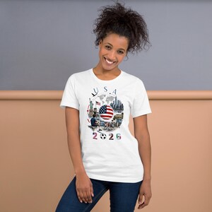 Pu&ograve; includere: T-shirt bianca con un design grafico colorato. Il design include la scritta "USA", la bandiera americana, un pallone da calcio e il numero "2026". La maglietta &egrave; realizzata in un materiale morbido.