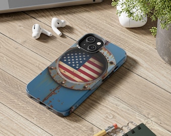 Può includere: Una custodia blu per telefono con un design a bandiera americana invecchiata. La bandiera è inserita in un cerchio di metallo arrugginito, aggiungendo un'estetica vintage. La custodia è su una superficie di legno, vicino a un laptop, auricolari e una piccola pianta.