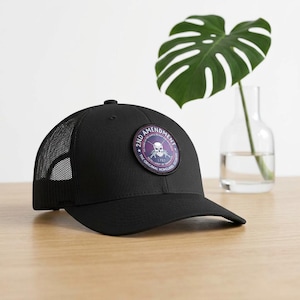 May include: A black trucker cap with a mesh back and a circular patch on the front. The patch features a skull and crossed rifles graphic with the text 2ND AMENDMENT, THE ORIGINAL HOMELAND SECURITY, and 1789 in a purple and white colour scheme.