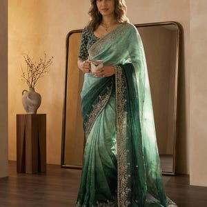 Puede incluir: Una mujer con un sari verde degradado con intrincados bordados en el borde. El sari pasa de verde claro a oscuro. Sostiene una taza y está de pie frente a un espejo. El sari es de kamalkriti by mona.