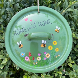Könnte beinhalten: Ein runder grüner Metalldeckel mit einem mittigen Griff und einer Juteschnur zum Aufhängen. Die Oberfläche zeigt den schwarzen Schriftzug "HOME sweet HOME", zwei rosa Herzen, drei gezeichnete Bienen und bunte Wildblumen.