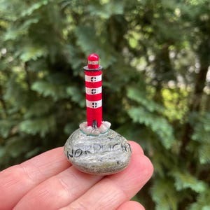 Könnte beinhalten: Eine kleine, handbemalte Leuchtturm-Figur mit einem rot-weiß gestreiften Turm, einem schwarzen Oberteil und einer roten Laterne. Der Leuchtturm ist auf einem glatten, grauen, ovalen Stein befestigt, auf dem das Wort NORDLICHT in schwarzer Tinte steht.