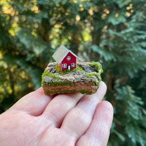 Könnte beinhalten: Ein Miniaturhaus in Rot mit silbernem Dach und weißen Fensterrahmen, befestigt auf einem Stück strukturierter brauner Baumrinde, die mit grünen Moosflecken bedeckt ist. Das kleine Bauwerk hat ein rundes Fenster und eine weiße Tür.