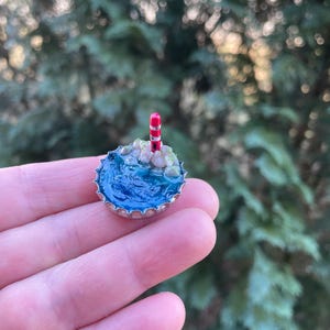 Könnte beinhalten: Eine Miniatur-Meereslandschaft, die in einem Flaschenverschluss gefertigt wurde. Die Szene zeigt einen blauen Ozean, kleine Felsen und einen rot-weiß gestreiften Leuchtturm. Der Flaschenverschluss hat einen silbernen Rand.