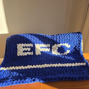 Könnte beinhalten: Eine blaue Strickdecke mit den Buchstaben "EFC" in Weiß. Ein weißer Streifen verläuft über die Decke. Die Decke ist über eine Holzoberfläche gefaltet.