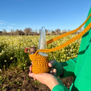 May include: A clear glass water pipe with a brown and gold mouthpiece is held in a mustard yellow crocheted cozy. The cozy has a matching strap. The background is a field of yellow flowers under a blue sky.