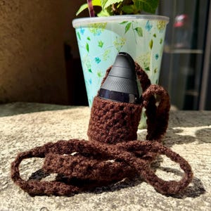 May include: A brown crocheted carrying case with a matching strap holds a black electronic device. The case is made of a textured yarn and is designed to hold the device securely. A light blue and white floral patterned pot with a green plant is in the background.