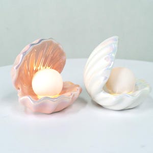 May include: Two seashell-shaped lamps with a pearlescent finish. One is pink, the other white. Each lamp holds a glowing, spherical light. The lamps are designed to resemble an open clam shell, creating a soft, ambient glow.