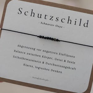 Könnte beinhalten: Ein dünnes schwarzes Schnurarmband mit sieben kleinen, facettierten schwarzen Onyxperlen in der Mitte. Das Armband ist auf einer rechteckigen weißen Präsentationskarte mit abgerundeten Ecken befestigt, die den Text Schutzschild und Schwarzer Onyx trägt.