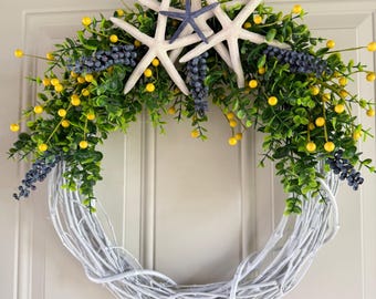 May include: A white twig wreath adorned with green foliage, yellow berries, and blue-grey clusters. Three white starfish and one blue starfish are centered at the top, creating a coastal theme. The wreath is suitable for a front door.