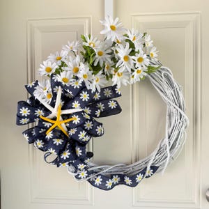May include: A white twig wreath decorated with white daisies, a navy blue ribbon with white daisy patterns, and two starfish. The wreath is hanging on a white door.