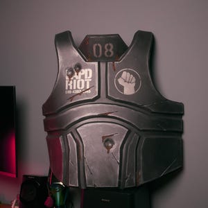 May include: A gray, weathered-looking chest armor piece with the text "PD RIOT" and the number "08". A white fist symbol is on the right side. The armor is displayed on top of a black speaker.