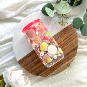May include: A clear glass tumbler with a pink lid, decorated with colorful illustrations of hearts, flowers, and cherries. The tumbler sits on a white and brown wooden serving board, with white flowers and greenery in the background.