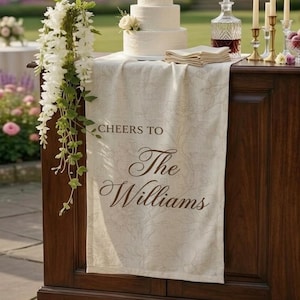May include: A three-tiered white wedding cake with floral accents sits atop a wooden cabinet. A cream-colored runner with the text "Cheers to The Williams" drapes over the cabinet. The scene includes candles, a decanter, and a bowl of fruit, set outdoors.