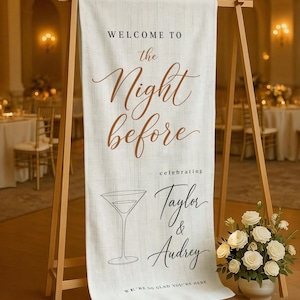 May include: A cream-colored welcome sign on a wooden easel. The sign reads "Welcome to the Night before" in elegant script, celebrating Taylor & Audrey. A martini glass illustration is below the text, with a floral arrangement in a white pot.