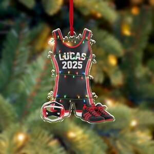 May include: A wrestling-themed Christmas ornament. The ornament is shaped like a wrestling singlet, with the name "LUCAS" and the year "2025" printed on it. It is black with red trim, a wrestling helmet, and a pair of wrestling shoes. The ornament is decorated with small lights.