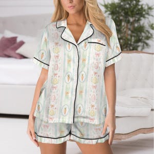 May include: A two-piece pajama set featuring a white background with vertical stripes in light blue and green. The pattern includes Easter eggs, carrots, and bows. The set has black trim and a button-down top with short sleeves.