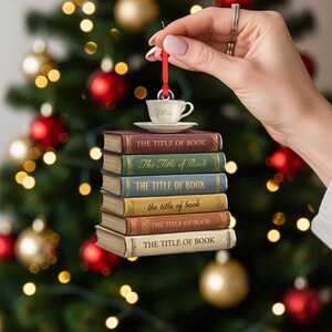 May include: A Christmas tree ornament shaped like a stack of books with a teacup on top. Each book is a different colour and has "THE TITLE OF BOOK" printed on the spine. The teacup is white and has the name "Mia" written on it.