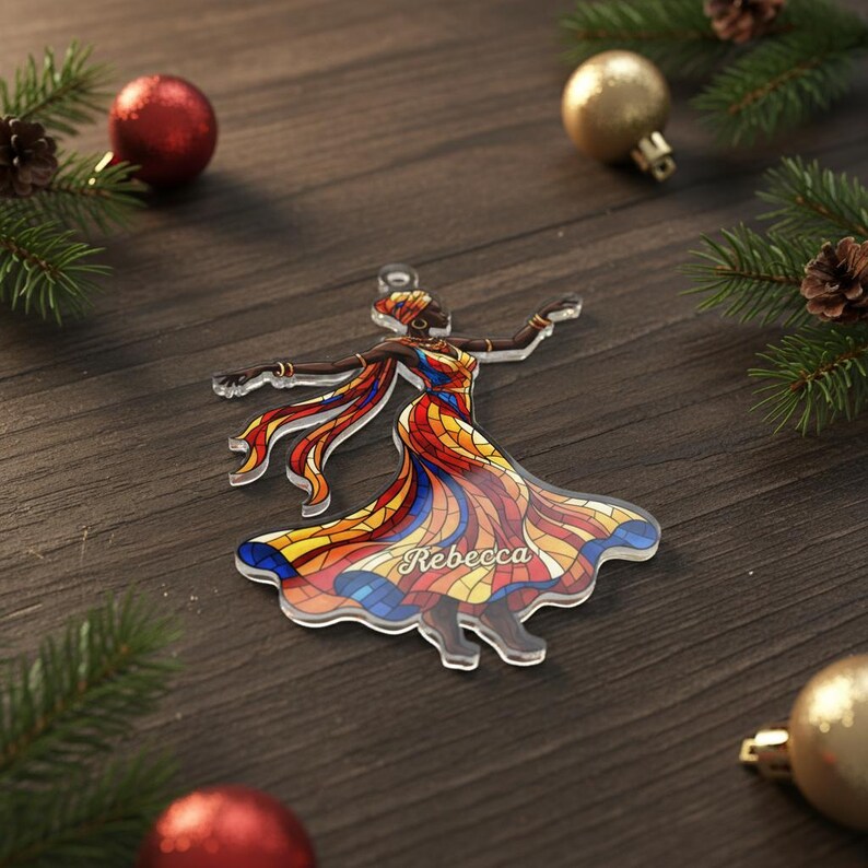 May include: A clear acrylic Christmas ornament depicting a woman in a vibrant, mosaic-style dress. The dress is a mix of red, yellow, and blue. The name "Rebecca" is visible. Festive decorations surround the ornament.