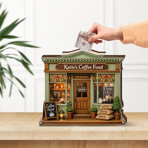 Op de afbeelding: Een decoratieve houten spaarpot in de vorm van een koffiehuis met de tekst "Katie's Coffee Fund". De spaarpot is geschilderd in groen en bruin, met een klein schoolbordje met "Coffee Fund".