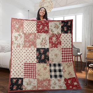 May include: A patchwork quilt with squares of floral, heart, and gingham patterns in red, black, and cream. The quilt has a red border and is held up by a person.