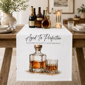 May include: A white table runner with the text "Aged To Perfection" and "Welcome to Scott's 40th Birthday." The runner features a watercolor illustration of a whiskey decanter and a rocks glass. Various bottles of liquor are arranged on the runner.
