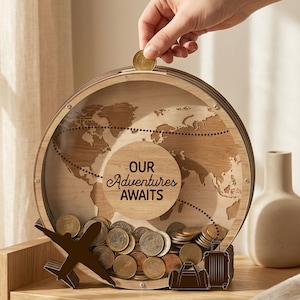 Op de afbeelding: Een houten, wereldbolvormige spaarpot met de tekst "OUR Adventures AWAITS". De spaarpot heeft een doorzichtige voorkant, een gleuf voor munten en een verzameling munten binnenin. Een hand laat een munt in de gleuf vallen. Inclusief een decoratief vliegtuig en bagage.