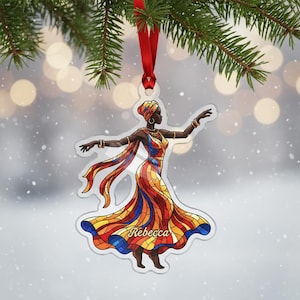 May include: A clear acrylic Christmas ornament depicting a woman in a vibrant dress, with the name "Rebecca". The ornament hangs from a red ribbon, set against a blurred backdrop of lights and fir needles.