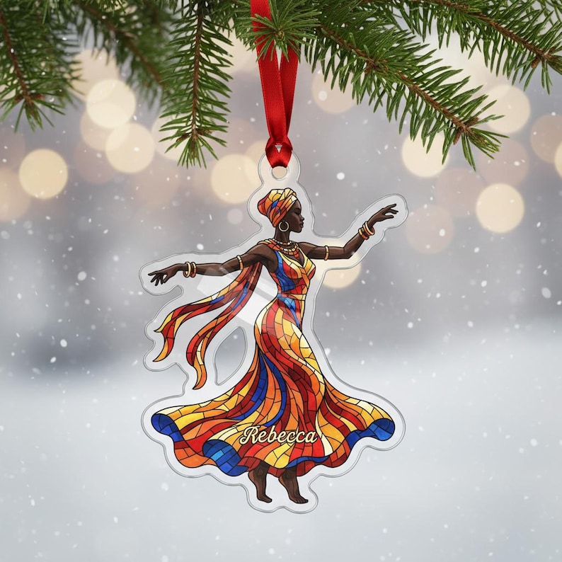 May include: A clear acrylic Christmas ornament depicting a woman in a vibrant dress, with the name "Rebecca". The ornament hangs from a red ribbon, set against a blurred backdrop of lights and fir needles.