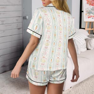 May include: A two-piece pajama set featuring a repeating pattern of Easter eggs, carrots, and bows on a white background with vertical stripes in light blue and green. The short-sleeved top and shorts have black trim.